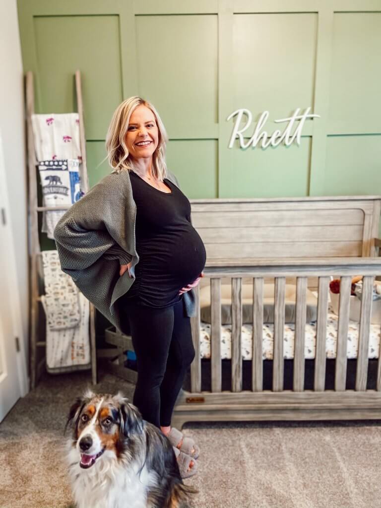 pregnant woman standing in front of crib with dog in frame; introducing your dog to your baby 