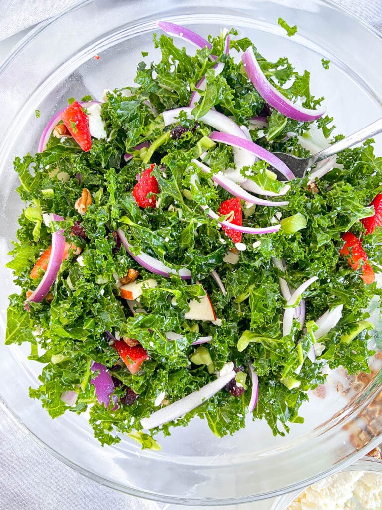 This is a photo of a large bowl of kale salad with mix-ins including chopped apples, red onions, strawberries, feta cheese and chopped pecans. 